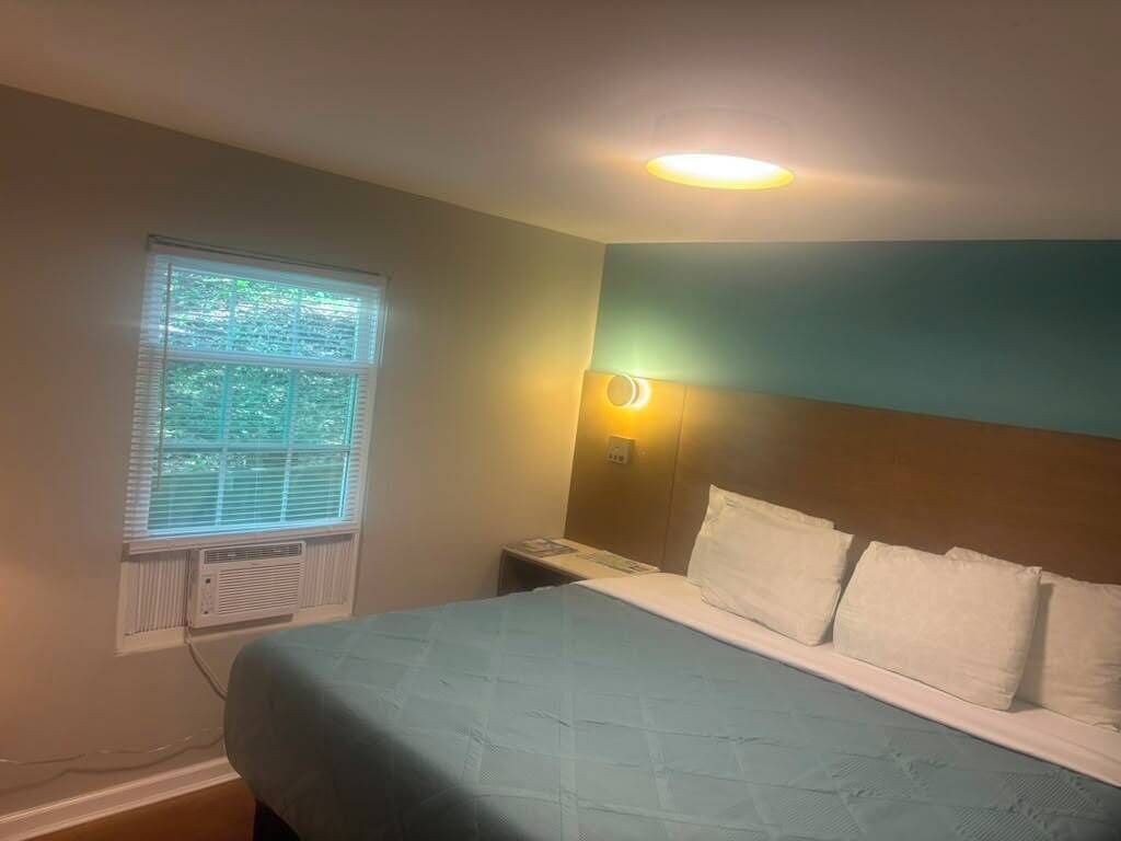 A bedroom with a bed, a window with blinds, a wall-mounted nightstand, and a ceiling light.