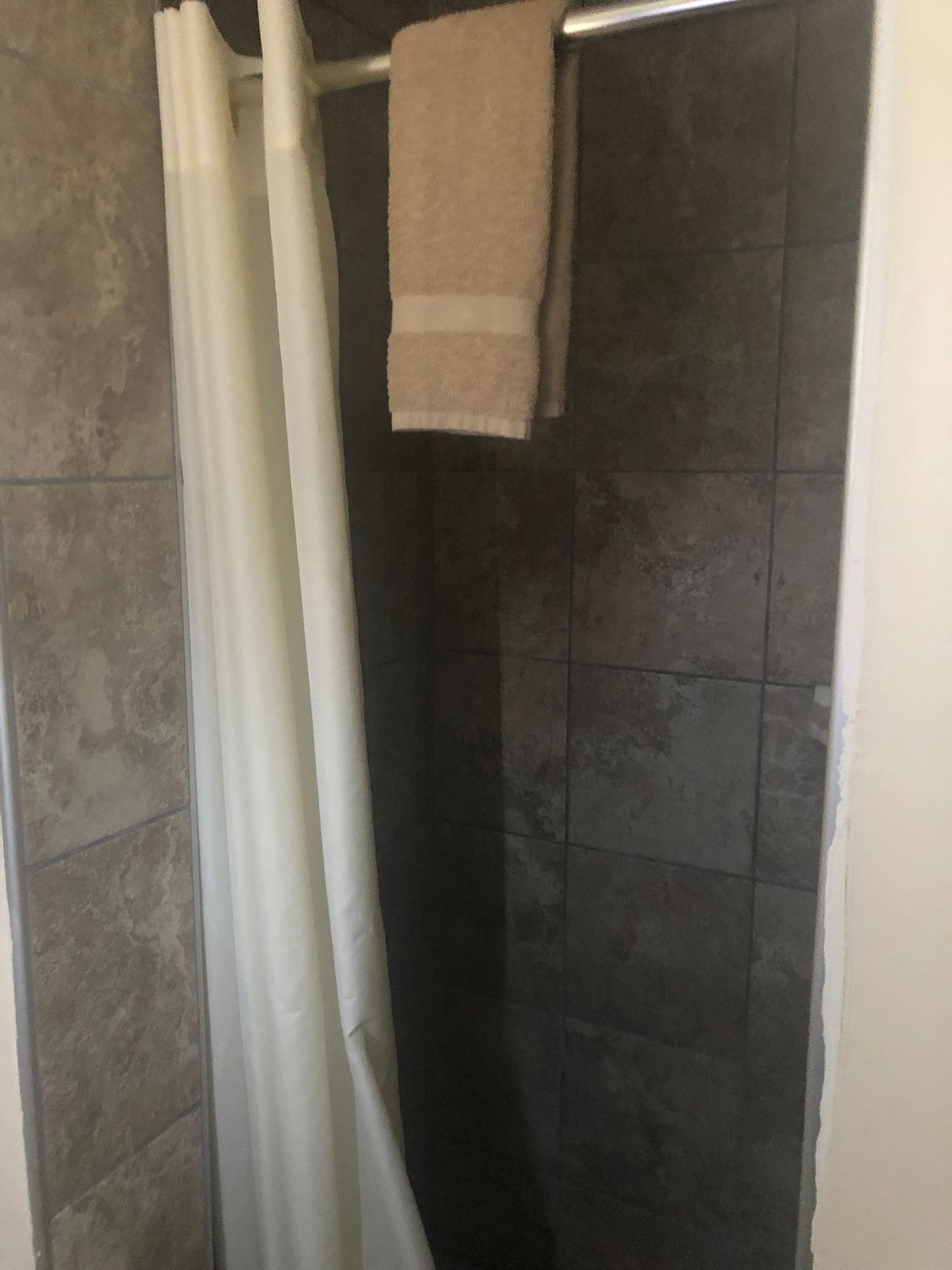 A shower area with dark tiled walls, a white curtain, and a beige towel hanging on a metal rod.