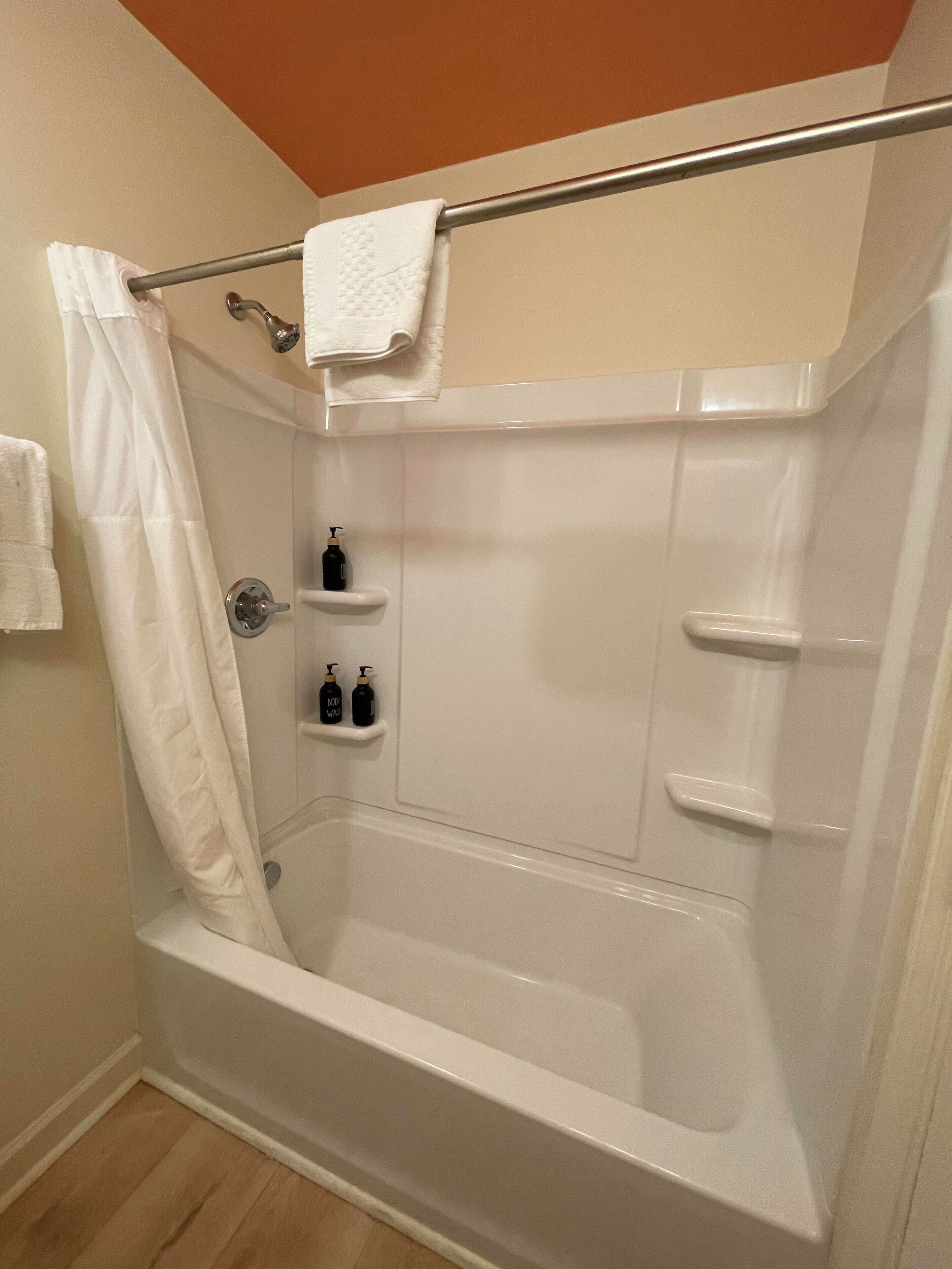 A corner shower with a white bathtub, a shower curtain, and two bottles of shampoo on a shelf.