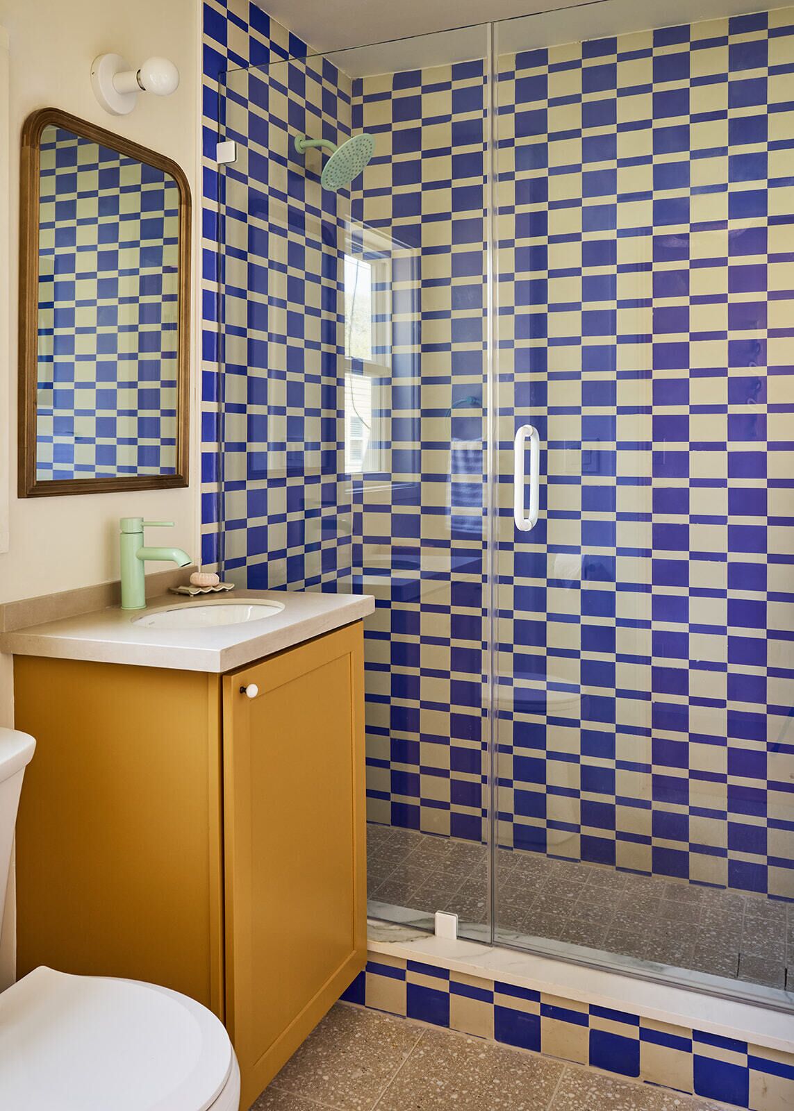 Ein Badezimmer mit Wandbelag aus blau-beigen Karokacheln, einer Glasdusche, einem weißen Waschbecken mit grünem Wasserhahn und einem Holzschrank.