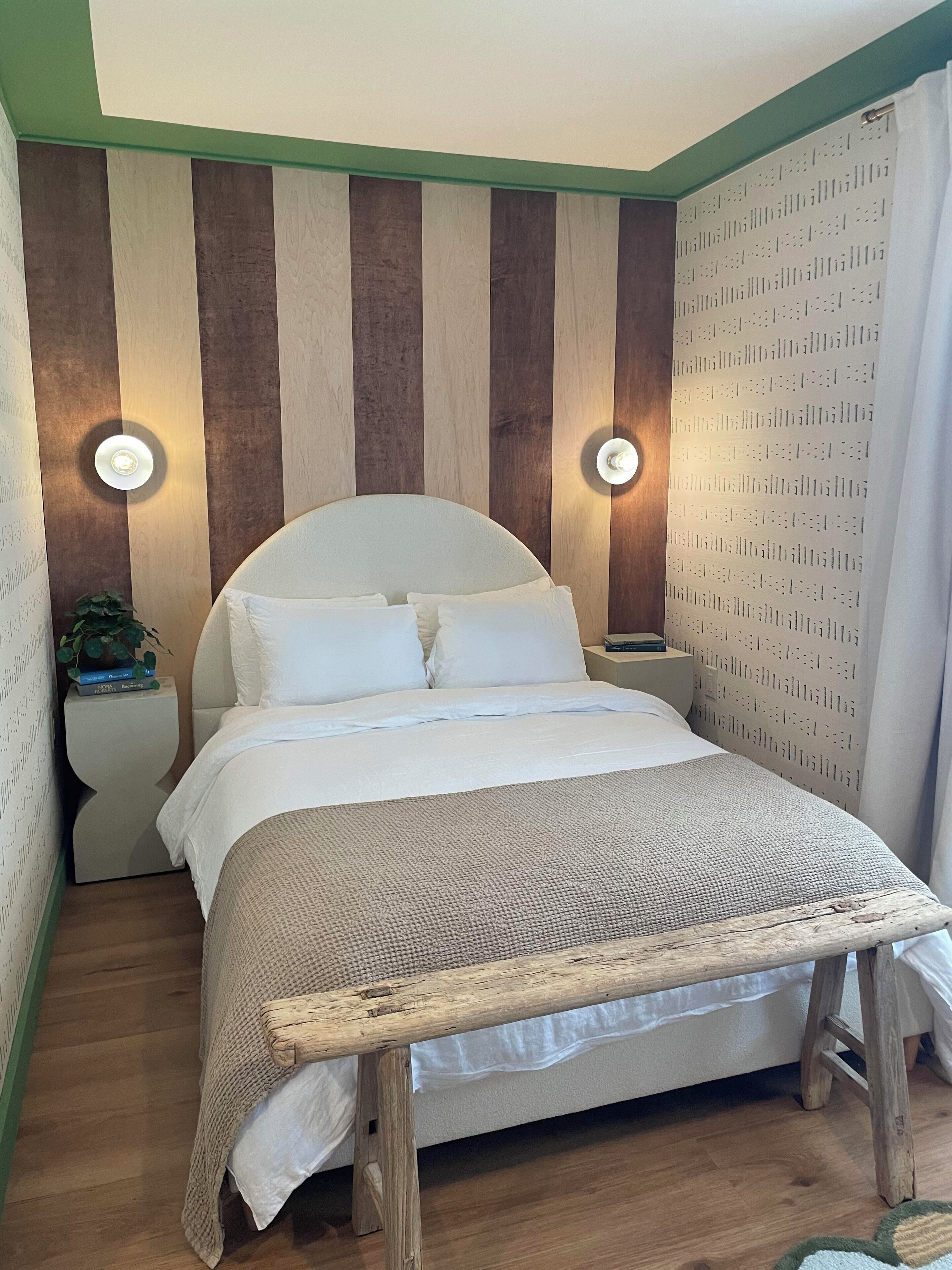A bed with a wooden headboard, a white mattress, and a woven footboard. There are two bedside tables with lamps, a potted plant, and a patterned wall.