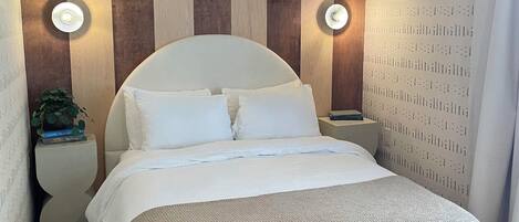 A bed with a wooden headboard, a white mattress, and a woven footboard. There are two bedside tables with lamps, a potted plant, and a patterned wall.
