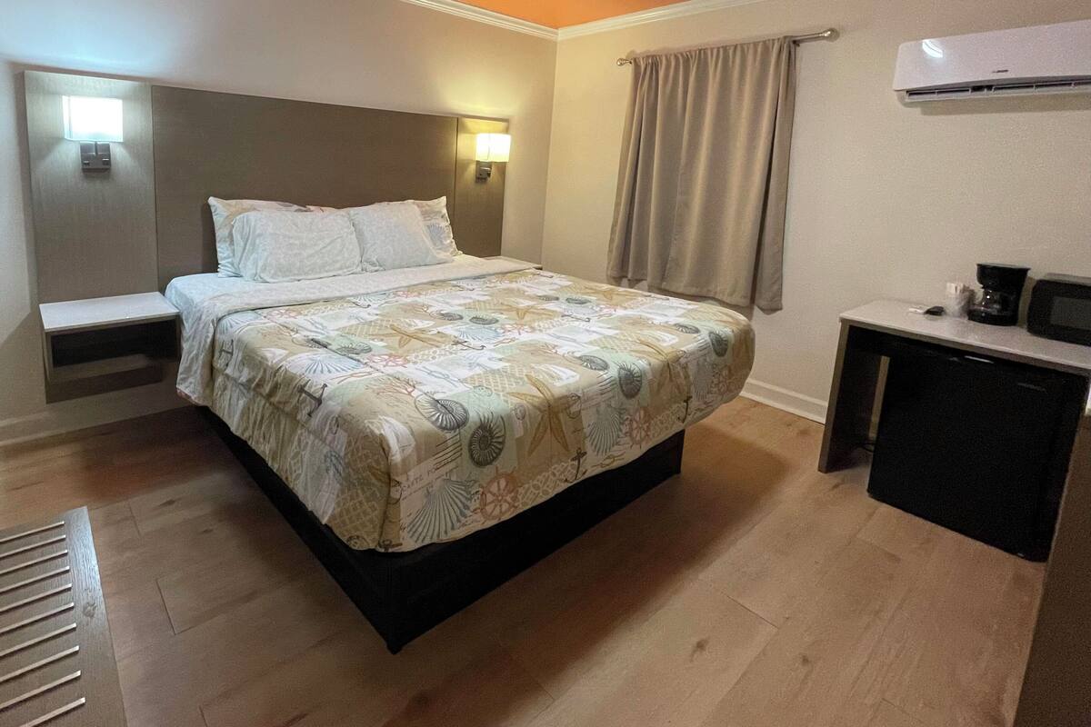 Ett hotellrum med en säng, ett sängbord, en lampa, en luftkonditioneringsanläggning och ett skrivbord med en stol.