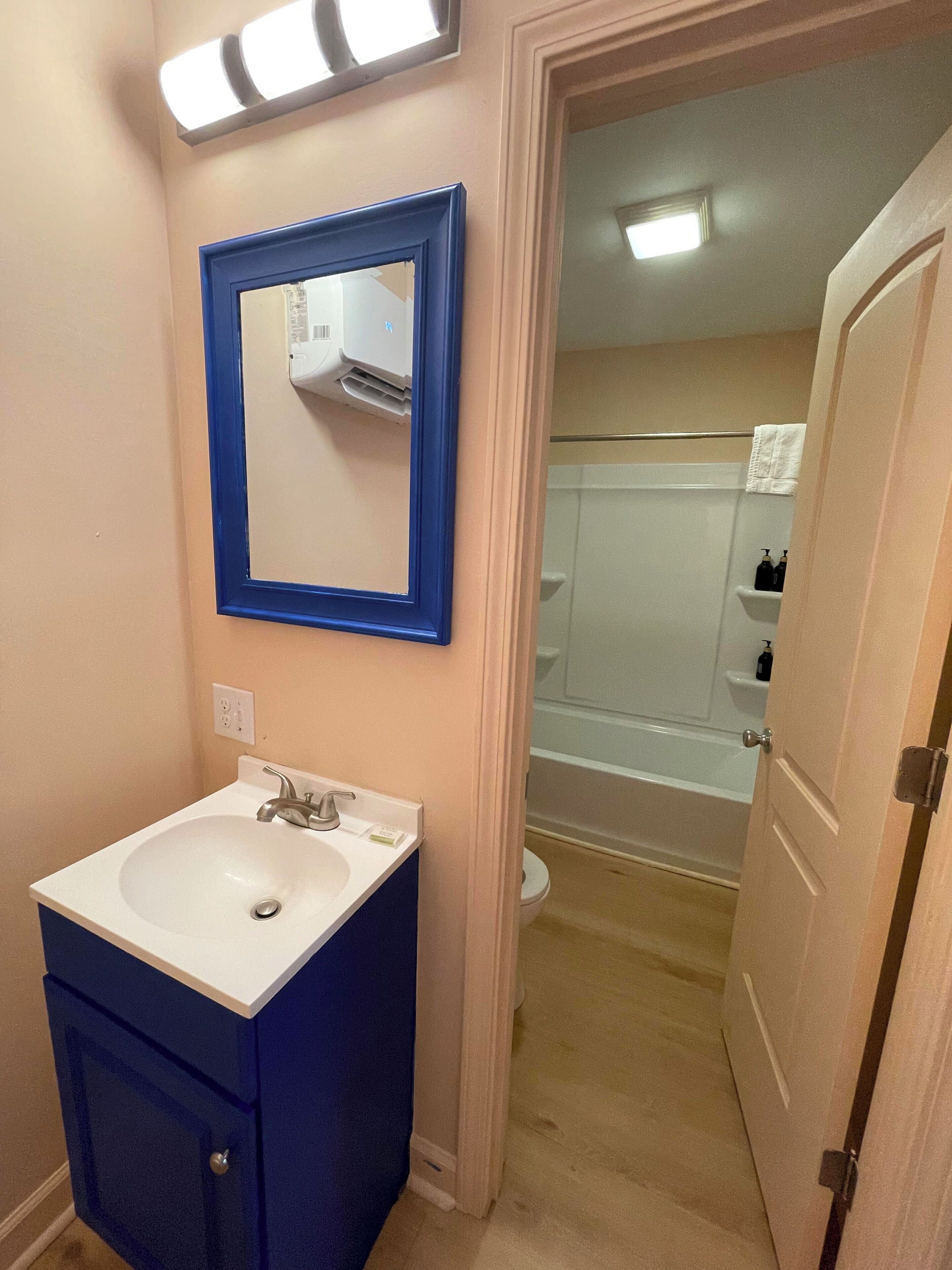 Une salle de bain avec un lavabo blanc, un meuble bleu et un miroir avec un cadre bleu. On aperçoit une douche au fond.