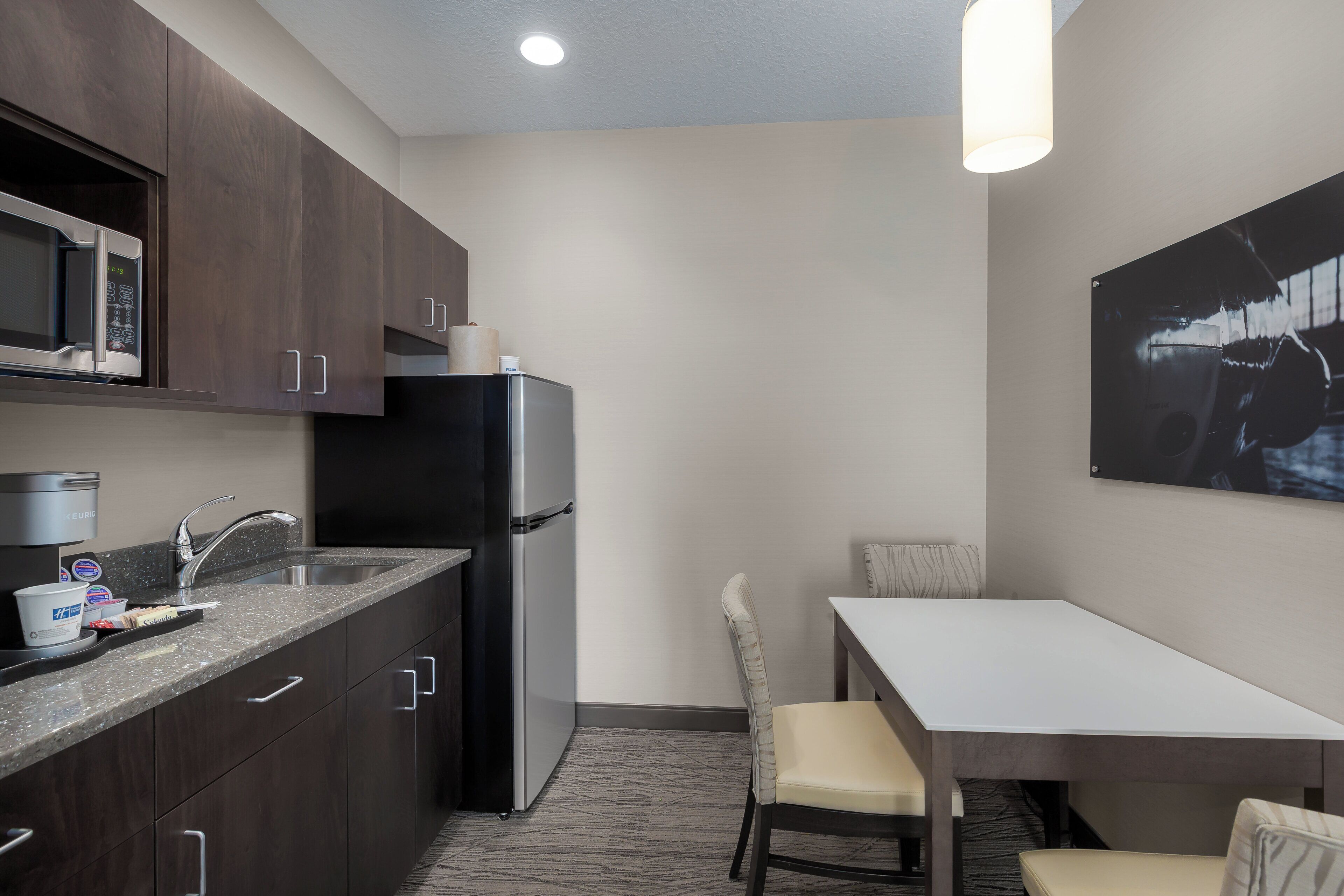 Una cocina compacta con muebles oscuros, un refrigerador de acero inoxidable, un microondas y una cafetera. Hay un pequeño comedor con una mesa y sillas.