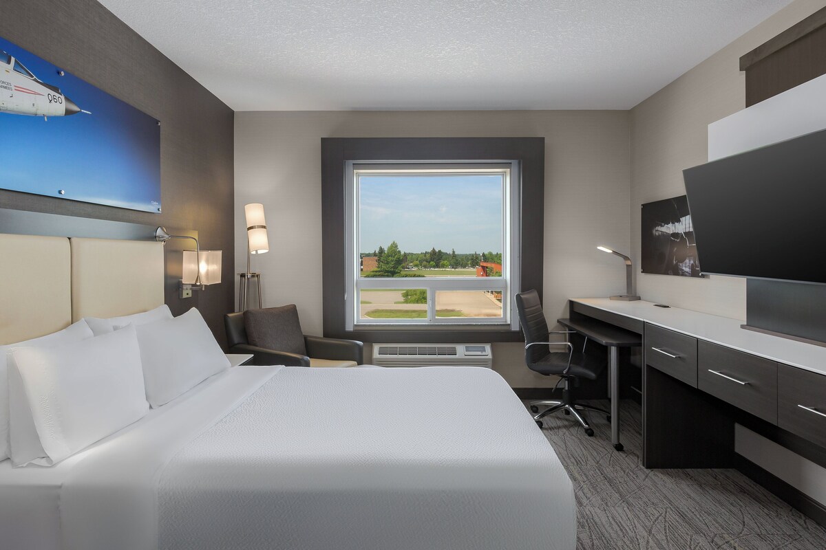 A hotel room with a large bed, a desk, a chair, a television, and a window with a view of a green landscape.