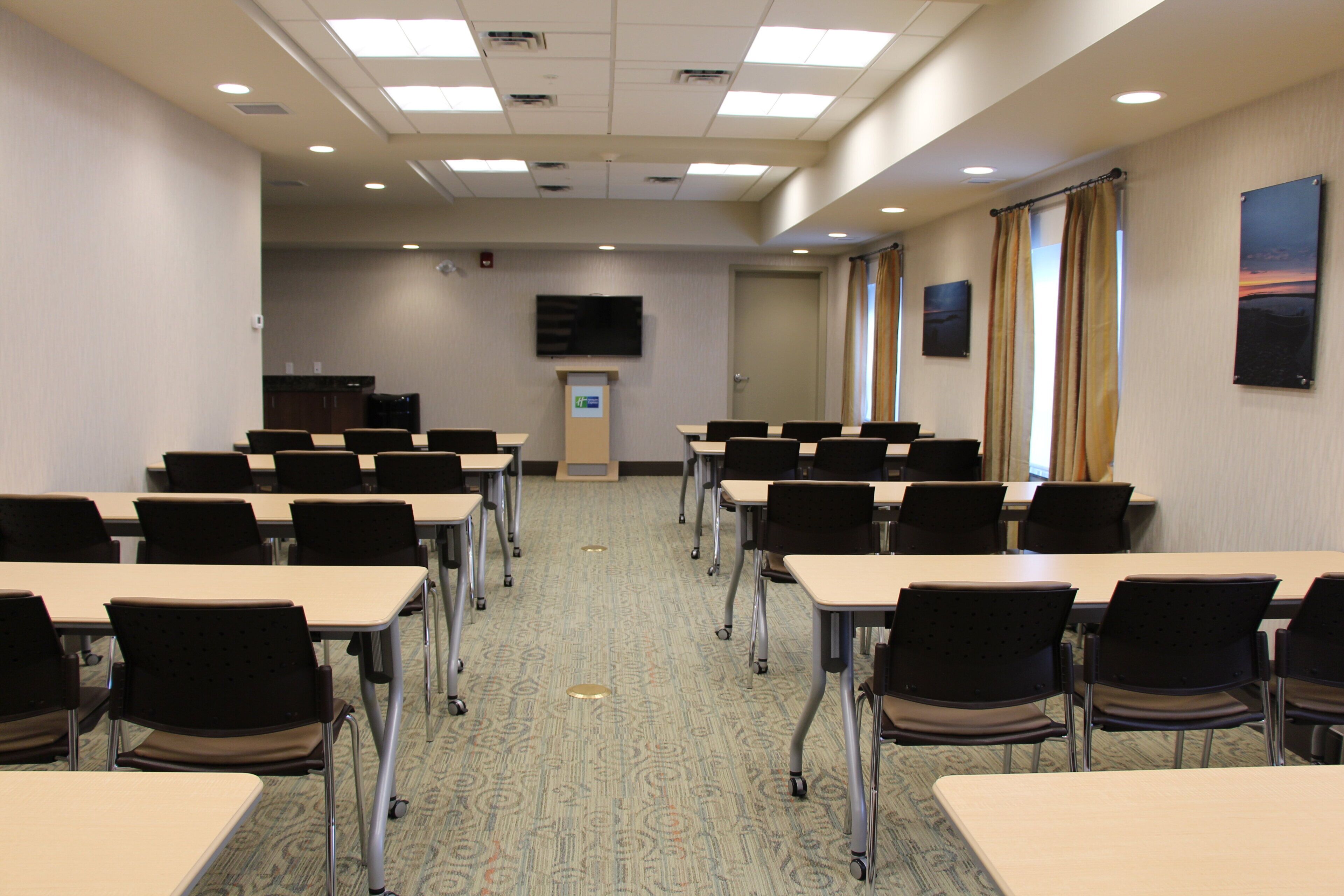 Un salón de conferencias con mesas y sillas dispuestas en filas, un atril, un televisor de pantalla plana y una puerta.