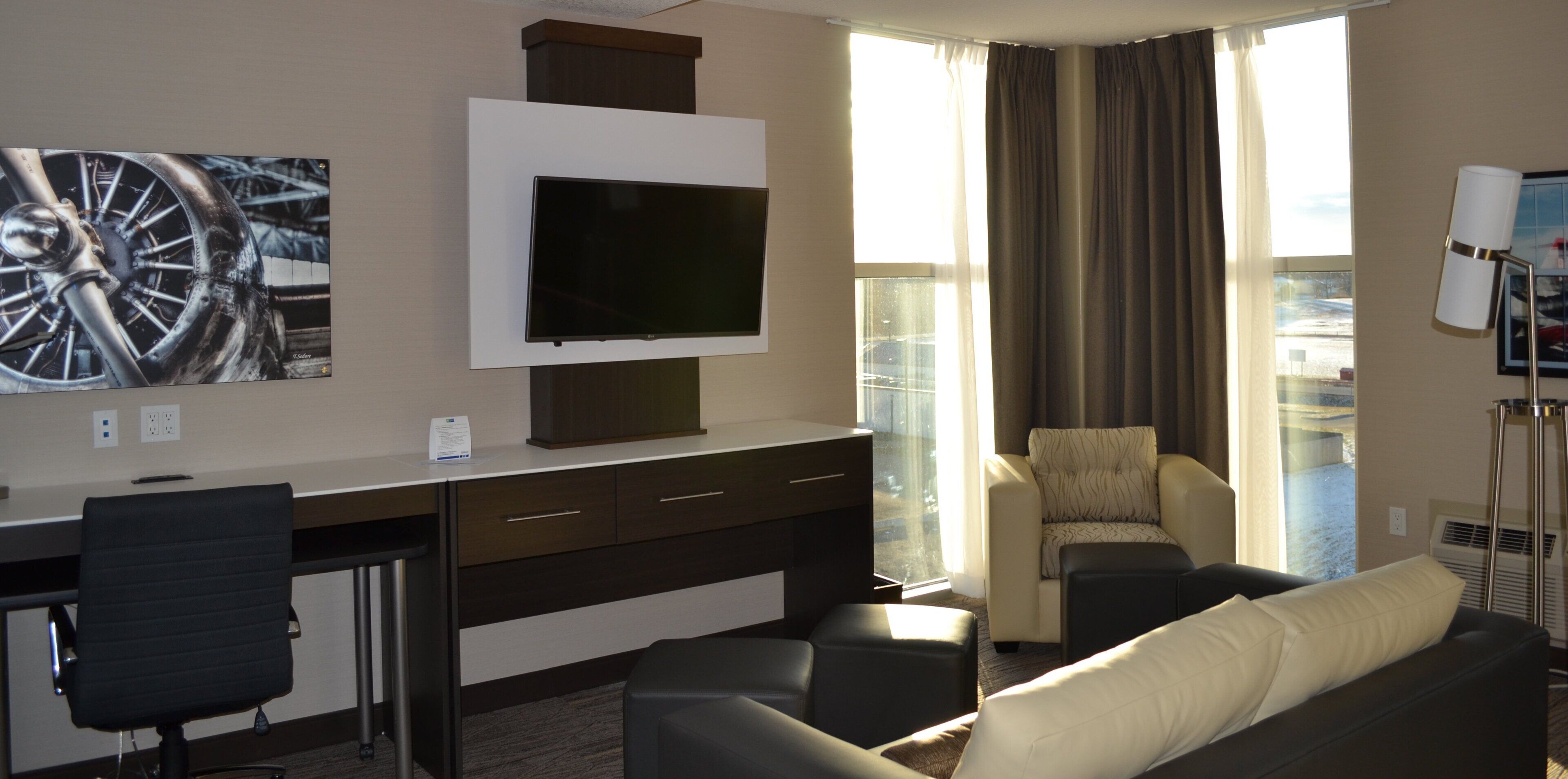 A hotel room with a flat-screen TV mounted on the wall, a desk with a chair, a sofa, and a window with curtains.