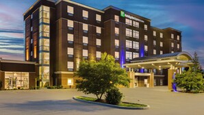 Um hotel Holiday Inn Express & Suites com design moderno, apresentando um amplo estacionamento e área ajardinada com árvores.