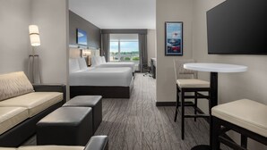 Quarto de hotel com uma cama grande, um sofá, uma escrivaninha e uma cadeira. Há uma janela com vista para a cidade.