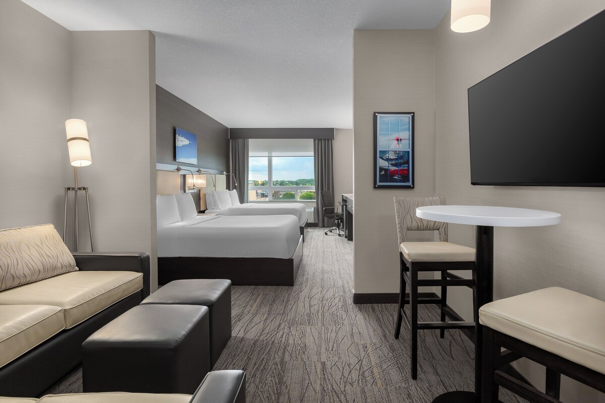 A hotel room with a large bed, a sofa, a desk, and a chair. There is a window with a view of the city.