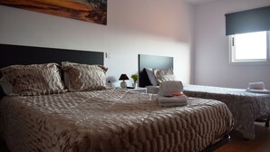 Un dormitorio con una cama grande, dos mesitas de noche, una mesita auxiliar, un cuadro y una ventana con cortina opaca.