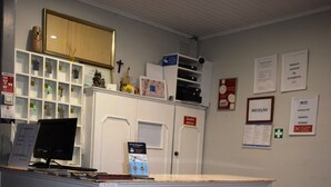 Mostrador de recepción con computadora, teléfono y una pequeña vitrina. Hay letreros en la pared, incluyendo uno que dice "RECEPCAO".