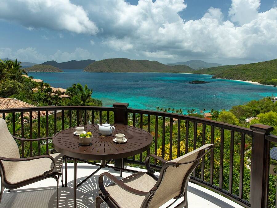 A balcony with a round table, two chairs, and a view of a turquoise sea and green hills.