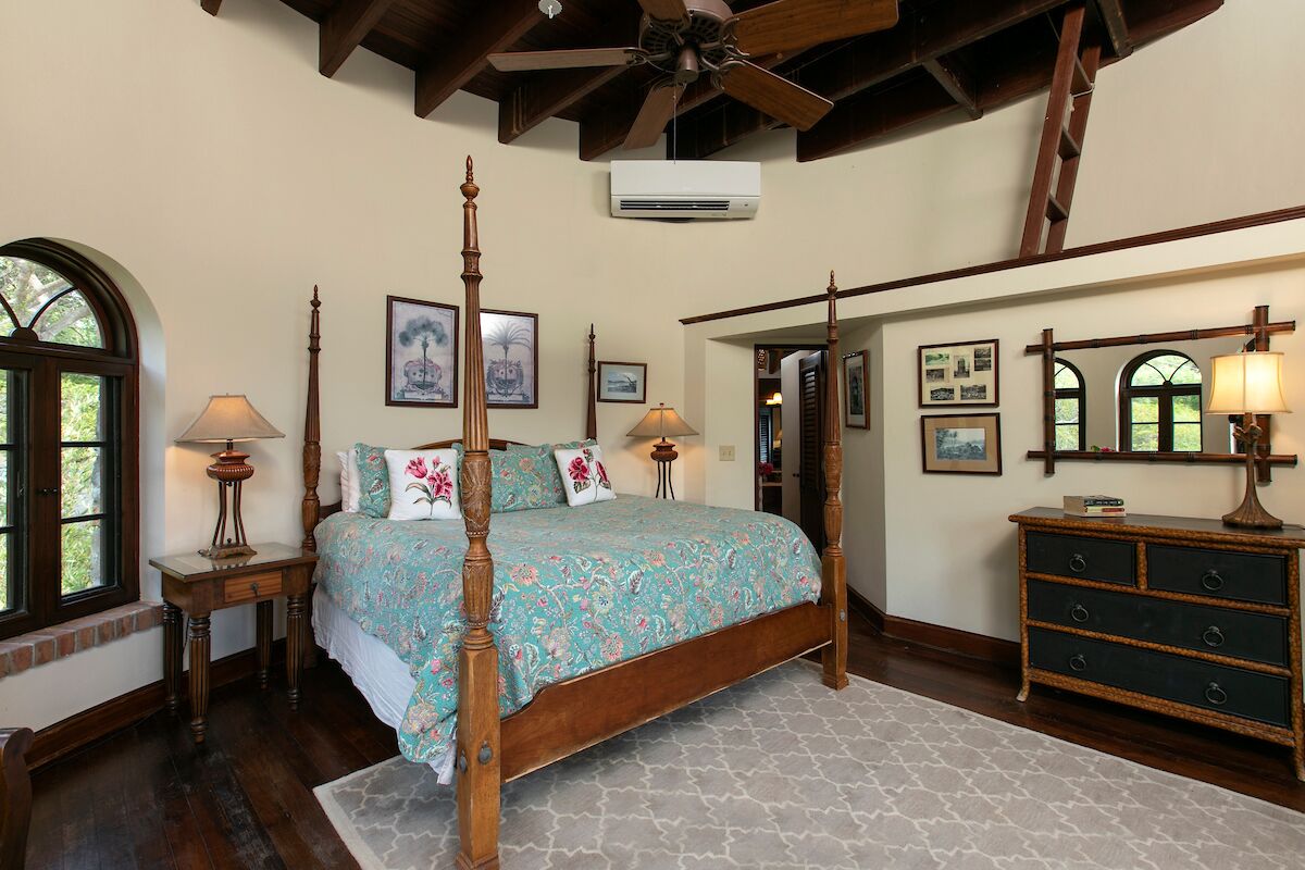 Un lit à baldaquin avec une couvre-lit à motifs floraux, une commode en bois avec des tiroirs et un ventilateur de plafond dans une pièce aux poutres apparentes et dotée d’une grande fenêtre.