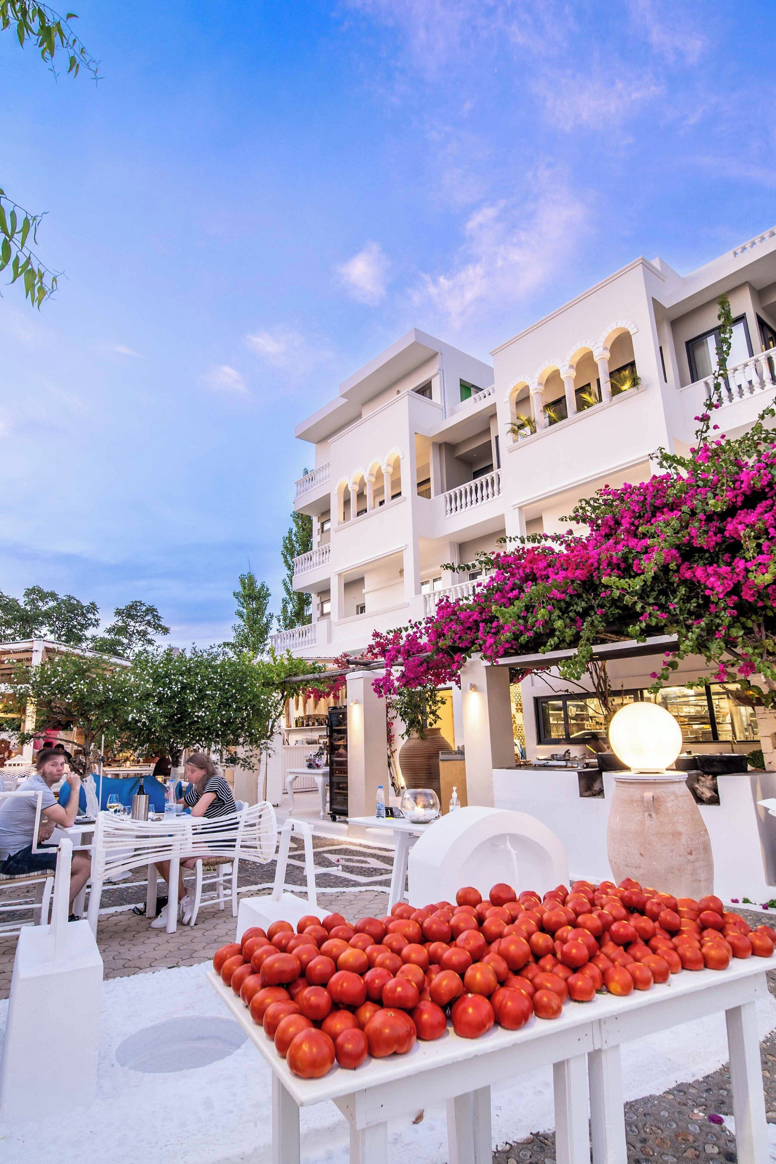 An outdoor seating area with white tables and chairs, a table of tomatoes, and a building with balconies and pink bougainvillea.