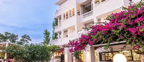 An outdoor seating area with white tables and chairs, a table of tomatoes, and a building with balconies and pink bougainvillea.