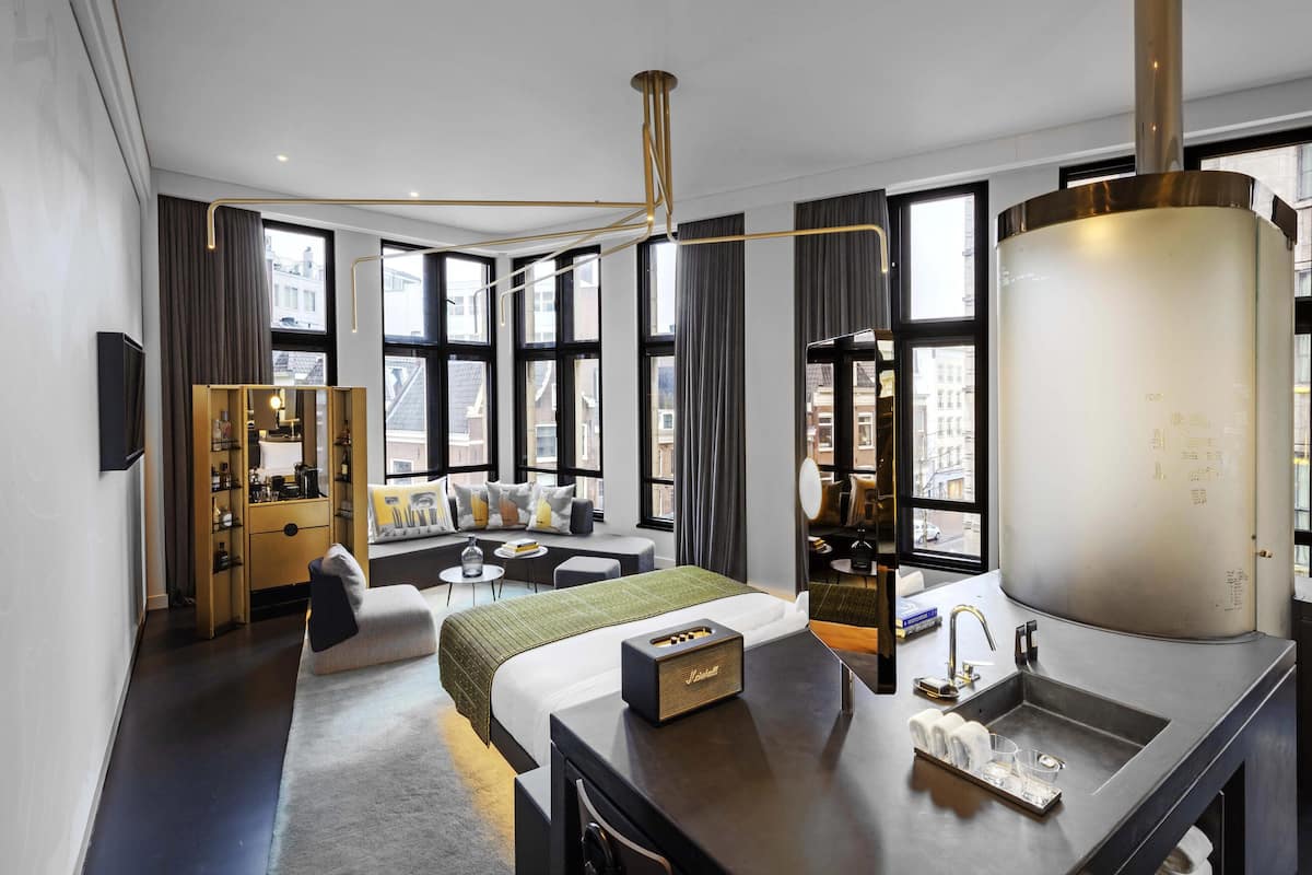 A modern hotel room with a large bed, a desk with a sink, a mirror, and a view of the city through large windows.