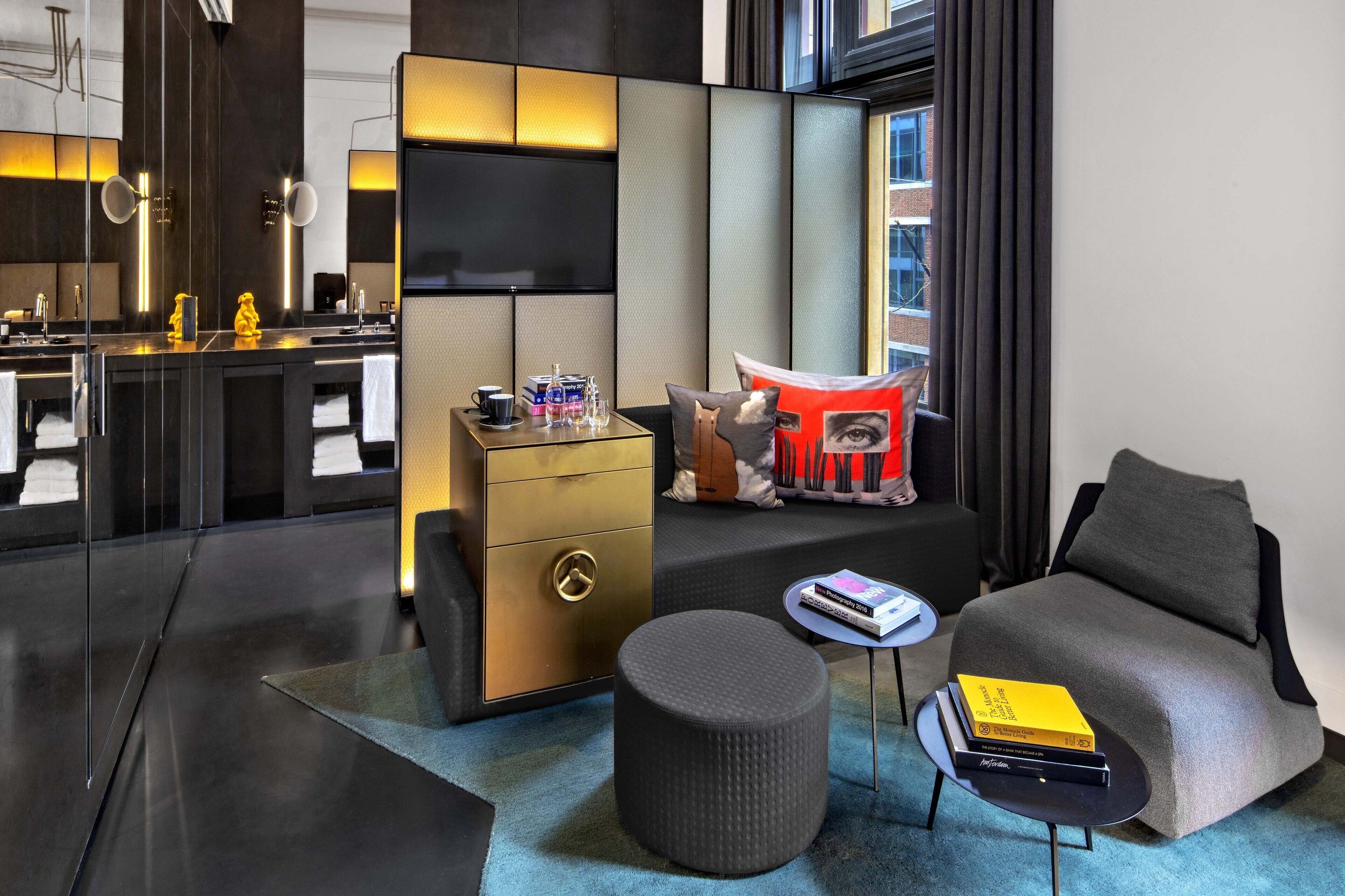 Habitación de hotel moderna con sofá, reposapiés y una mesita auxiliar con libros. Cuenta con un ventanal con cortinas, un televisor de pantalla plana en la pared y una nevera con distintos artículos.