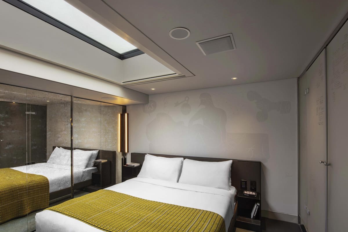 A modern hotel room with a large bed, a bedside table, a nightstand, a glass partition, and a skylight.