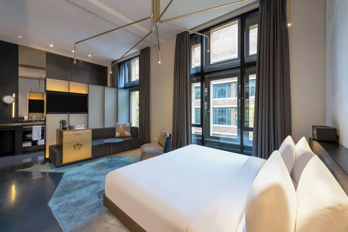 A modern hotel room with a large bed, a desk, a sofa, and a view of the city.