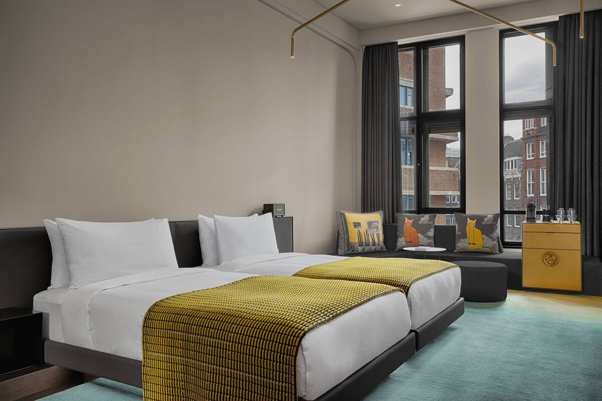 A modern hotel room with two beds, a sofa, a minibar, and large windows offering a city view.