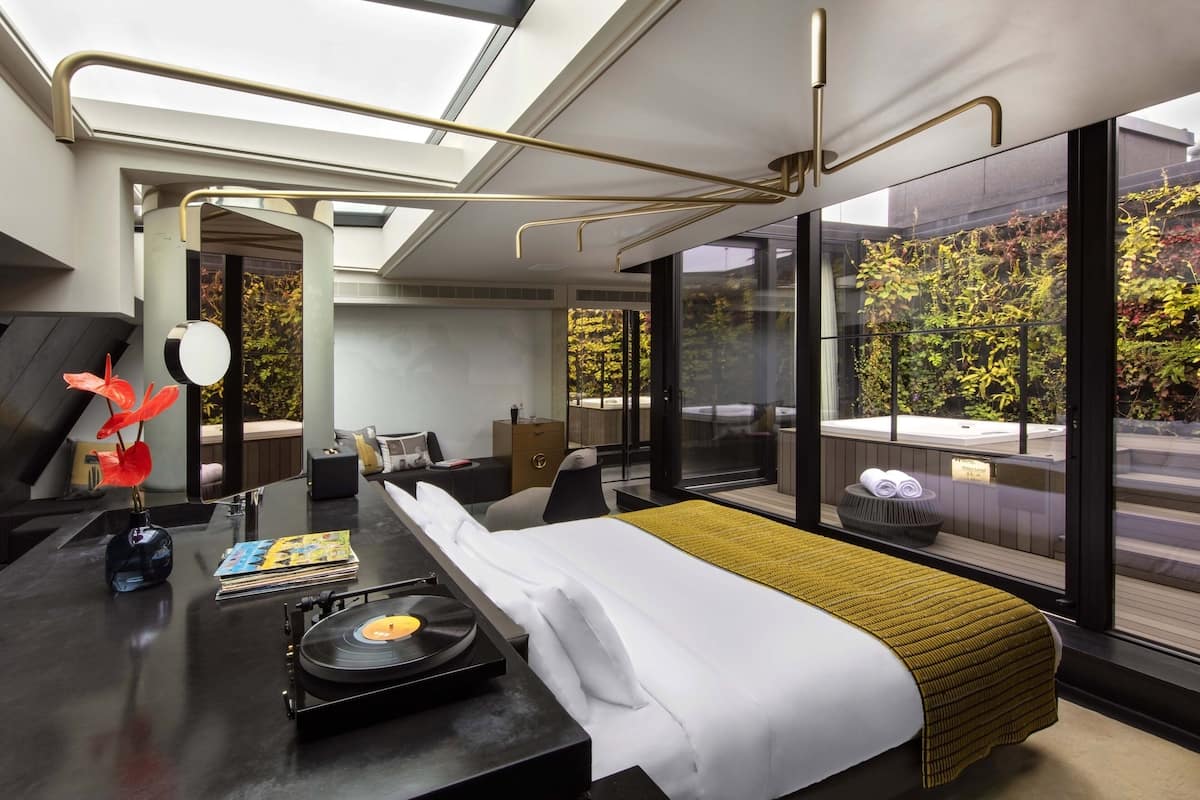 A modern hotel room with a large bed, a desk with a record player, a vase with flowers, and a view of trees through large windows.
