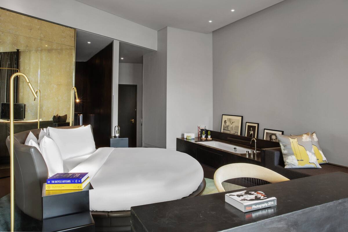 A modern hotel room with a large bed, a desk, a chair, a vanity with a mirror, and a large window.