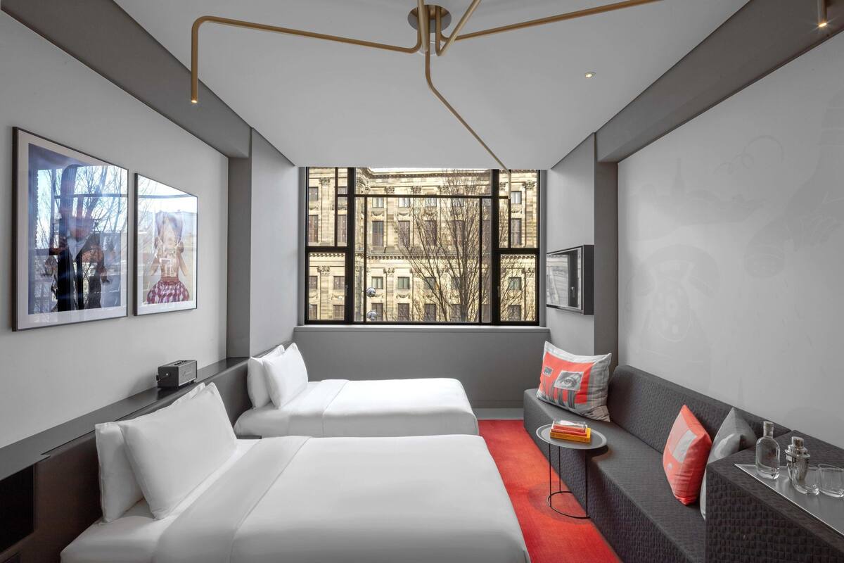 A modern hotel room with two beds, a sofa, a small table, and a large window offering a view of buildings.
