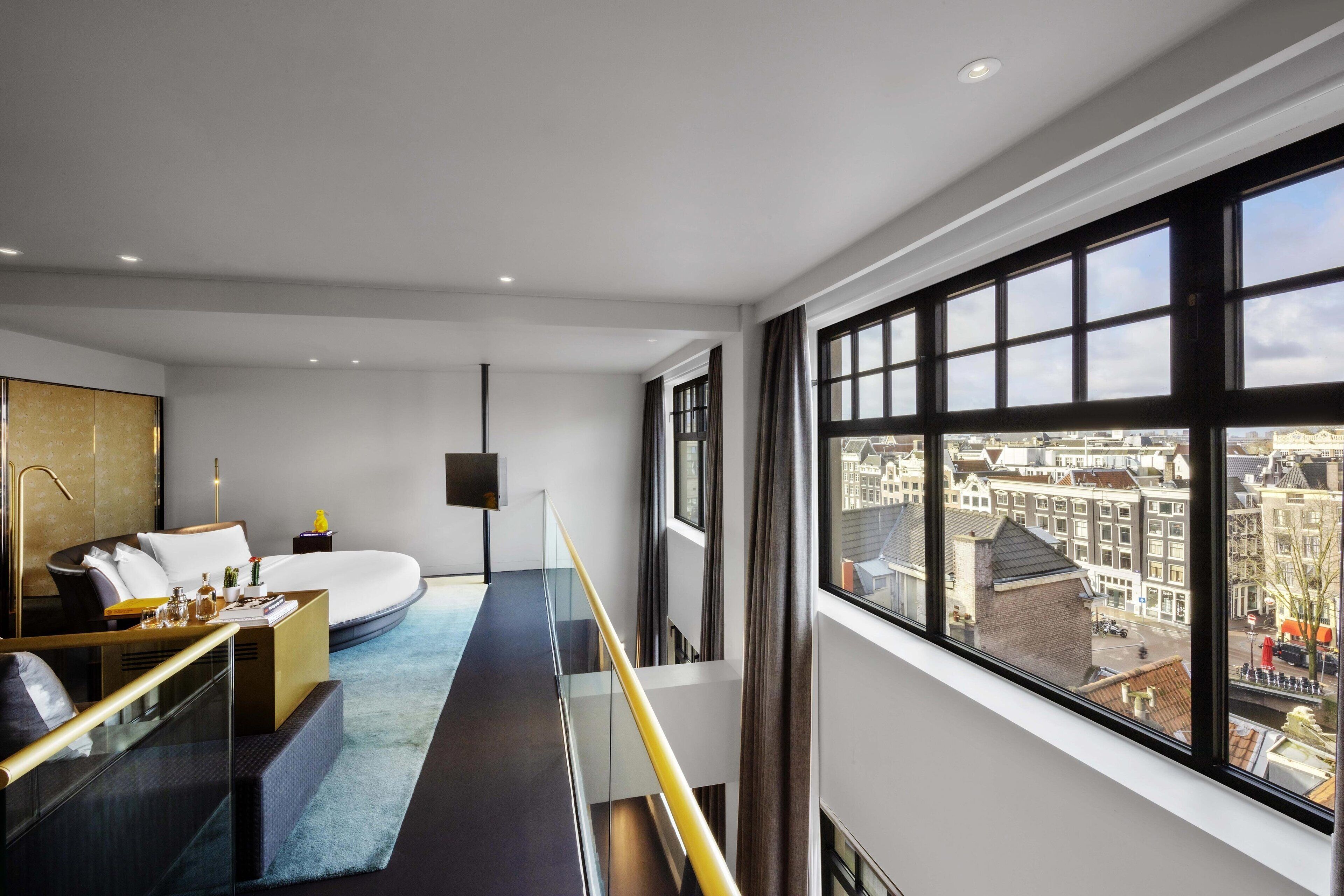 Habitación de hotel moderna con una cama grande, un área de descanso con barandilla de vidrio y vista a la ciudad a través de amplias ventanas.