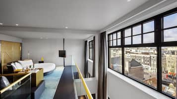 Habitación de hotel moderna con una cama grande, un área de descanso con barandilla de vidrio y vista a la ciudad a través de amplias ventanas.
