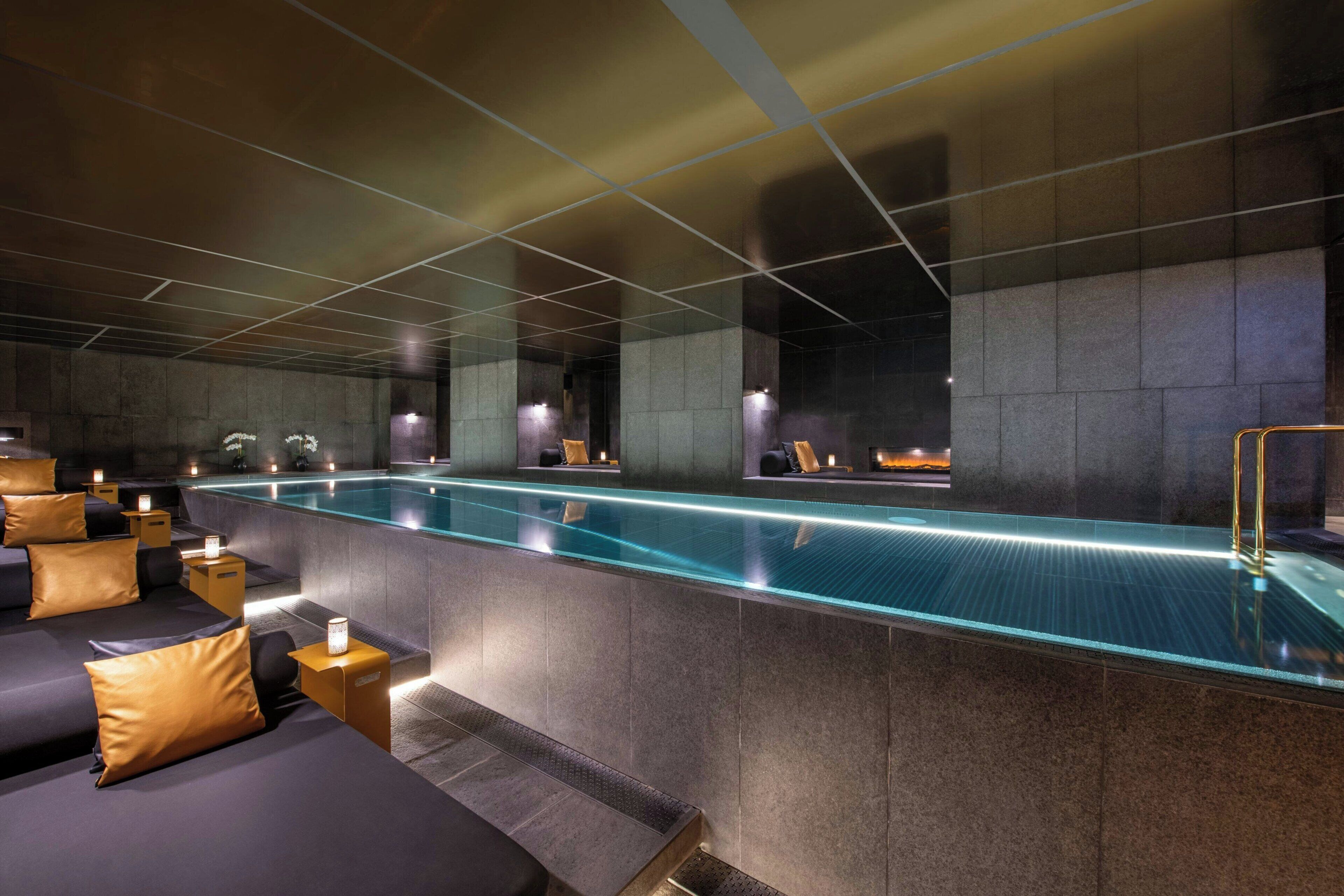 Un espace spa moderne doté d’une longue piscine rectangulaire, de coins salon avec des coussins et d’un éclairage d’ambiance.