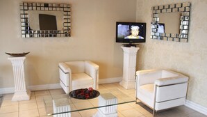 Uma sala de estar com dois poltronas brancas, uma mesa de centro de vidro, uma televisão e espelhos decorativos nas paredes.