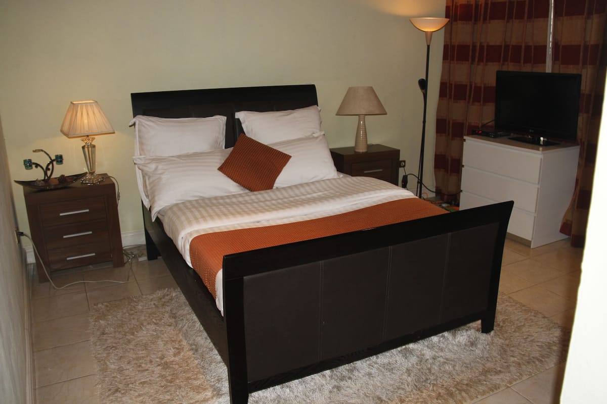 A neatly made bed with a brown throw blanket, two bedside tables with lamps, a television on a stand, and a dresser.