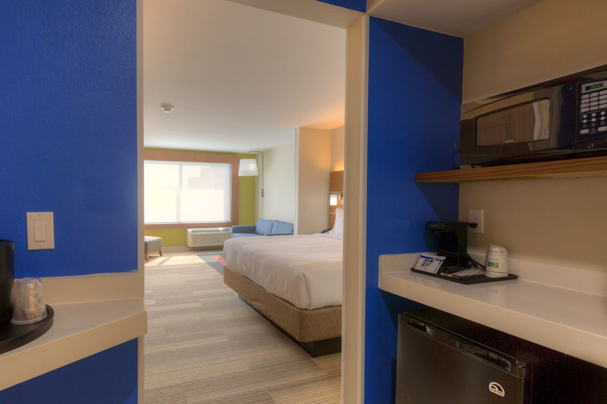 Hotellihuone, jossa on sänky, työpöytä, kahvinkeitin, mikroaaltouuni ja näkymä toiseen huoneeseen.