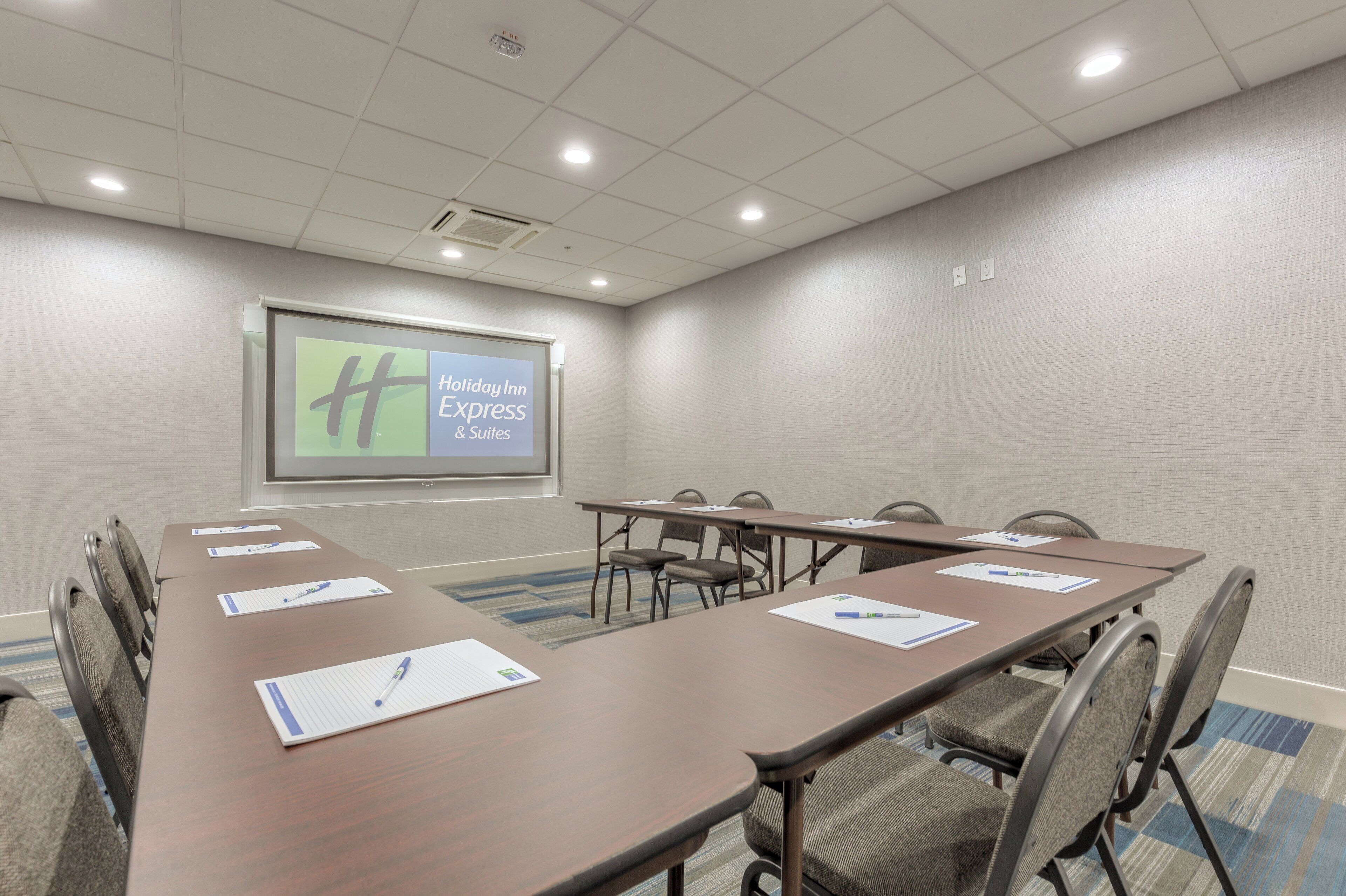 A conference room with a long table, chairs, a projector screen displaying 'Holiday Inn Express & Suites', and a fire alarm on the ceiling.
