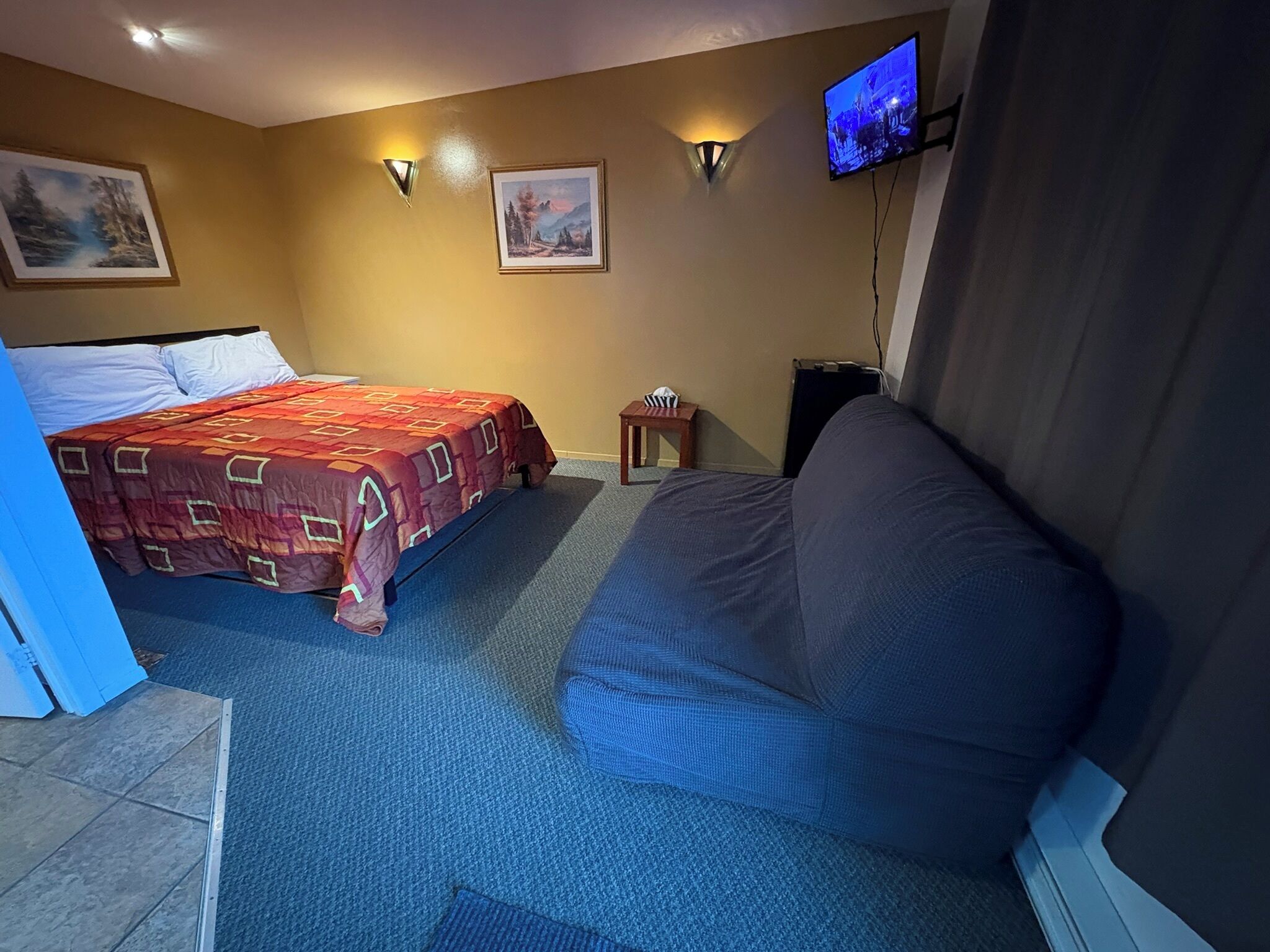 A hotel room with a bed, a sofa, a TV, and two wall-mounted lights.