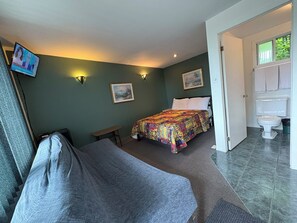Un dormitorio con cama, un banco, un cuadro y un baño visible a través de una puerta abierta.
