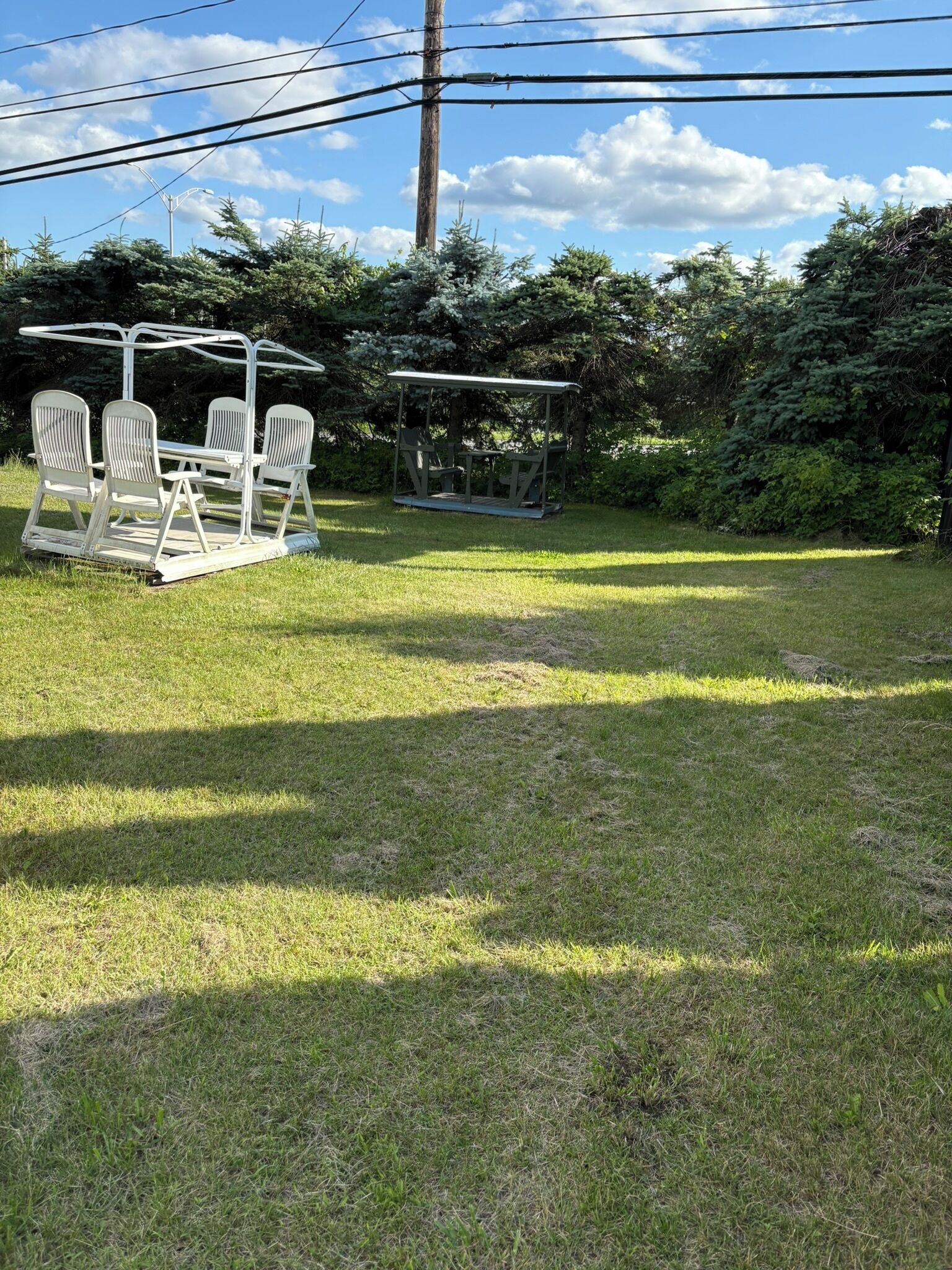 A grassy outdoor area with two white metal picnic tables and chairs, surrounded by trees and a utility pole.