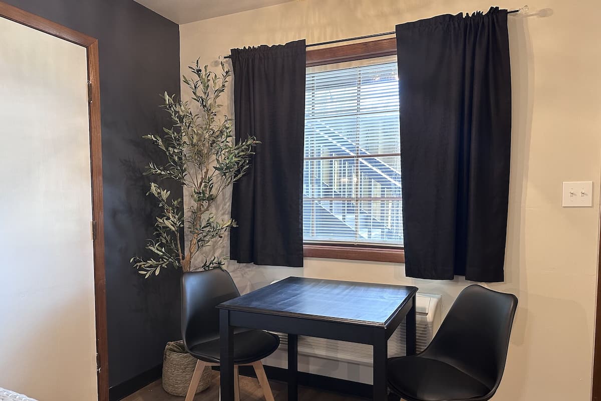 Een kleine kamer met een tafel, twee stoelen, een plant in pot en een raam met jaloezieën.