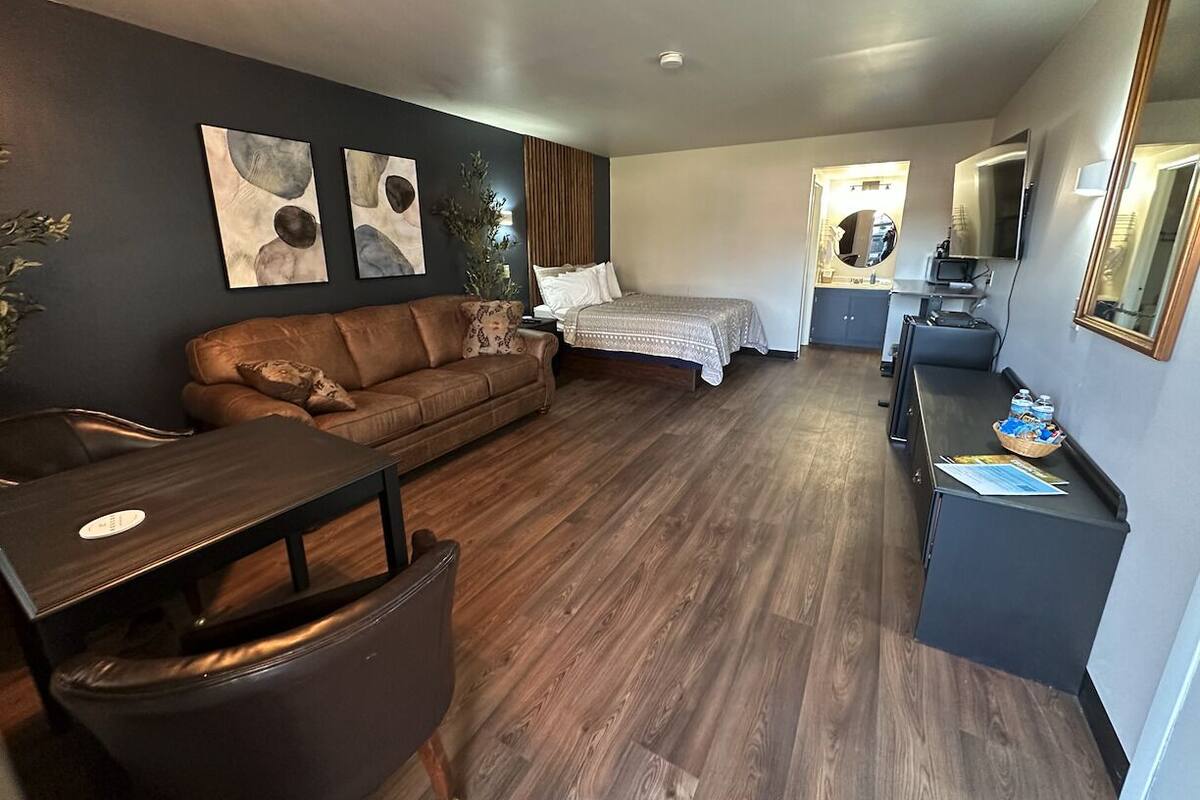 Een compacte hotelkamer met een bruine leren bank, een klein eettafeltje, een bed met een witte sprei, een bureau met een computer en een spiegel.