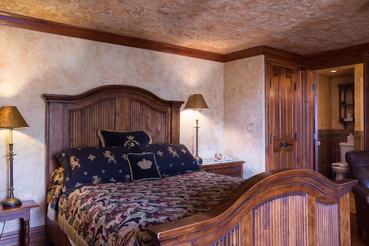 A bedroom with a large bed, wooden headboard, two bedside lamps, and a wooden door leading to a bathroom.