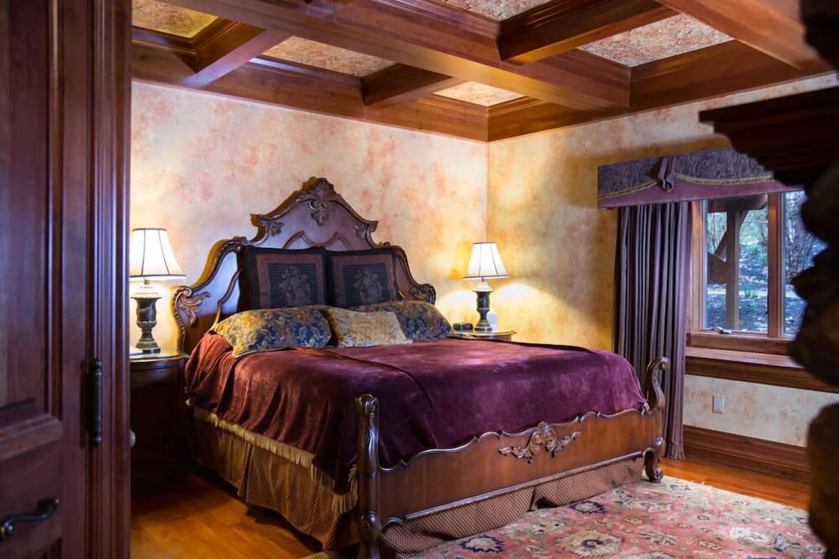 A traditional bedroom with a wooden bed, two bedside tables with lamps, a patterned rug, and a window with curtains.