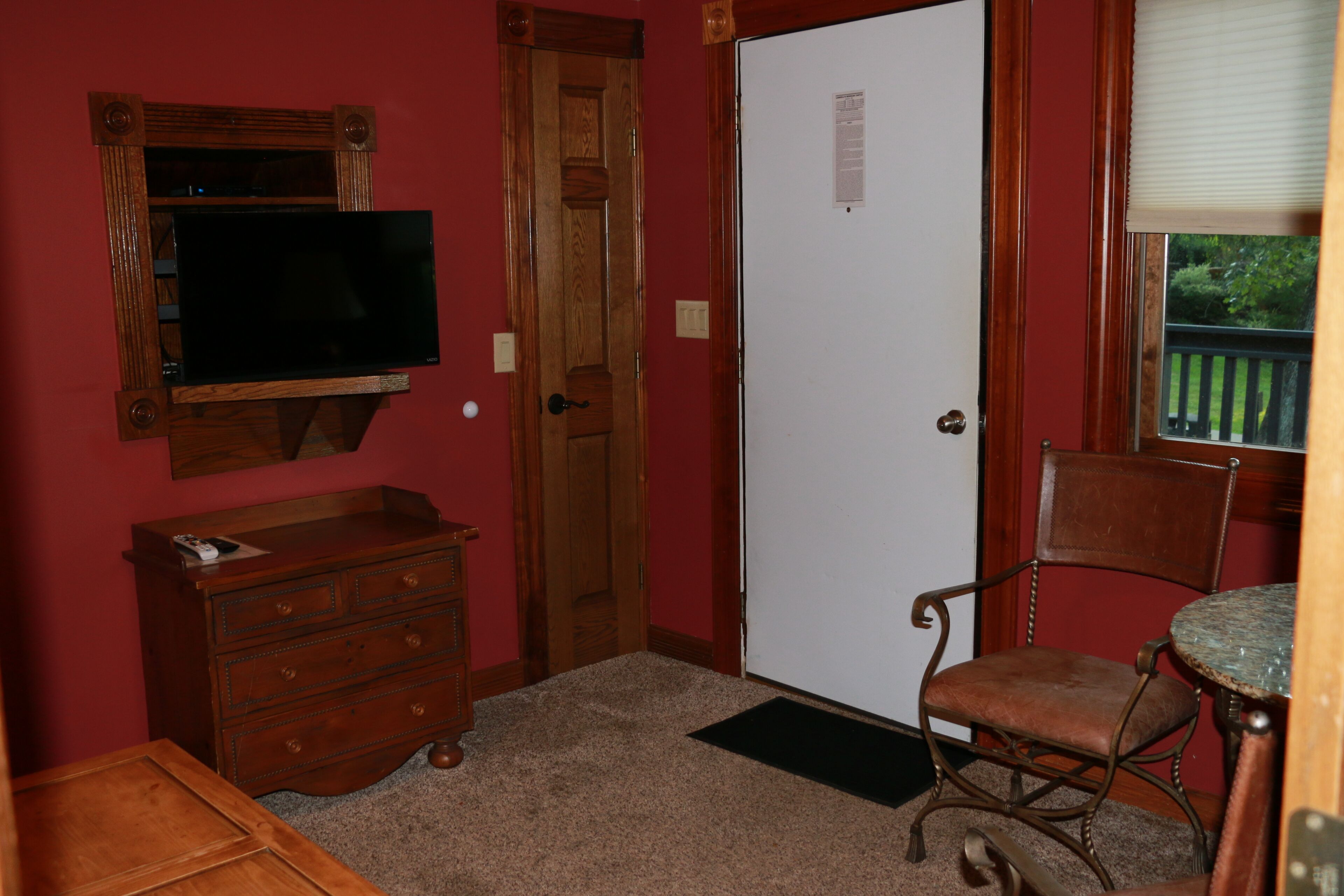 A room with a wooden dresser, a television mounted on the wall, a door, a chair, and a window with a view of greenery.