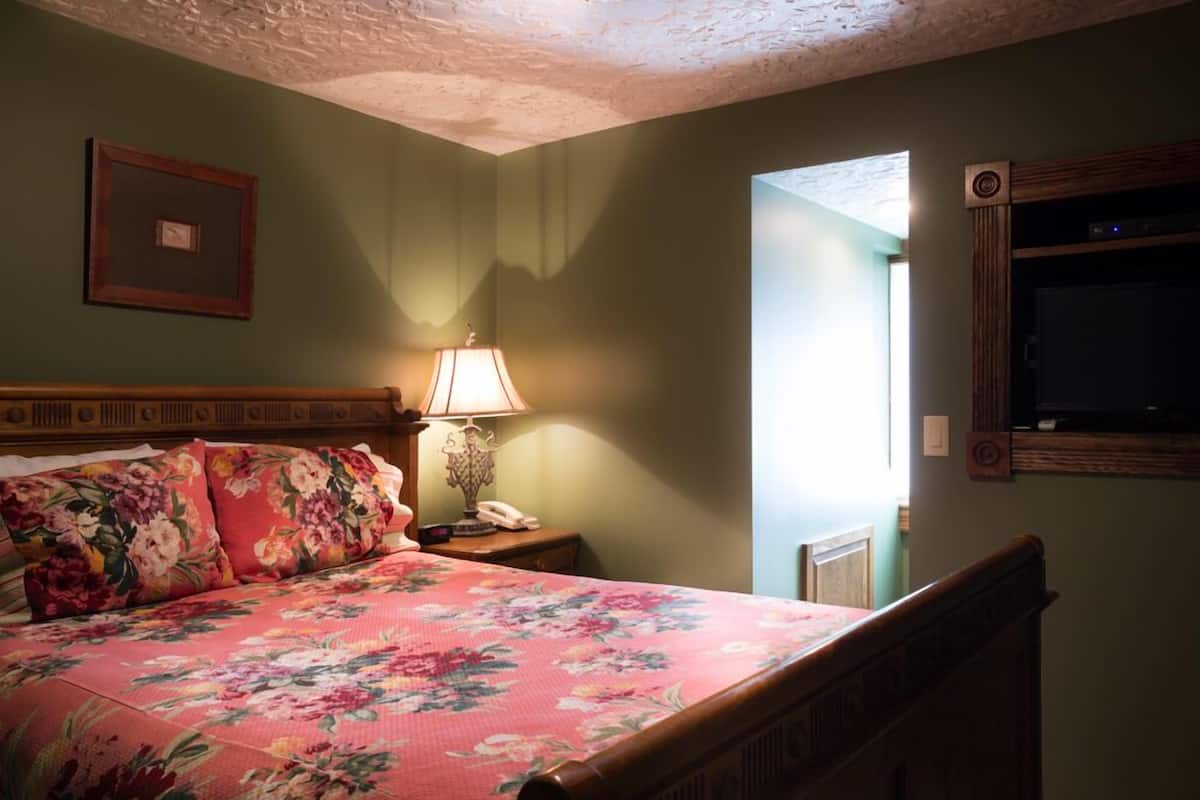 A bedroom with a floral bedspread, a bedside lamp, a framed picture on the wall, and a television mounted on the wall.