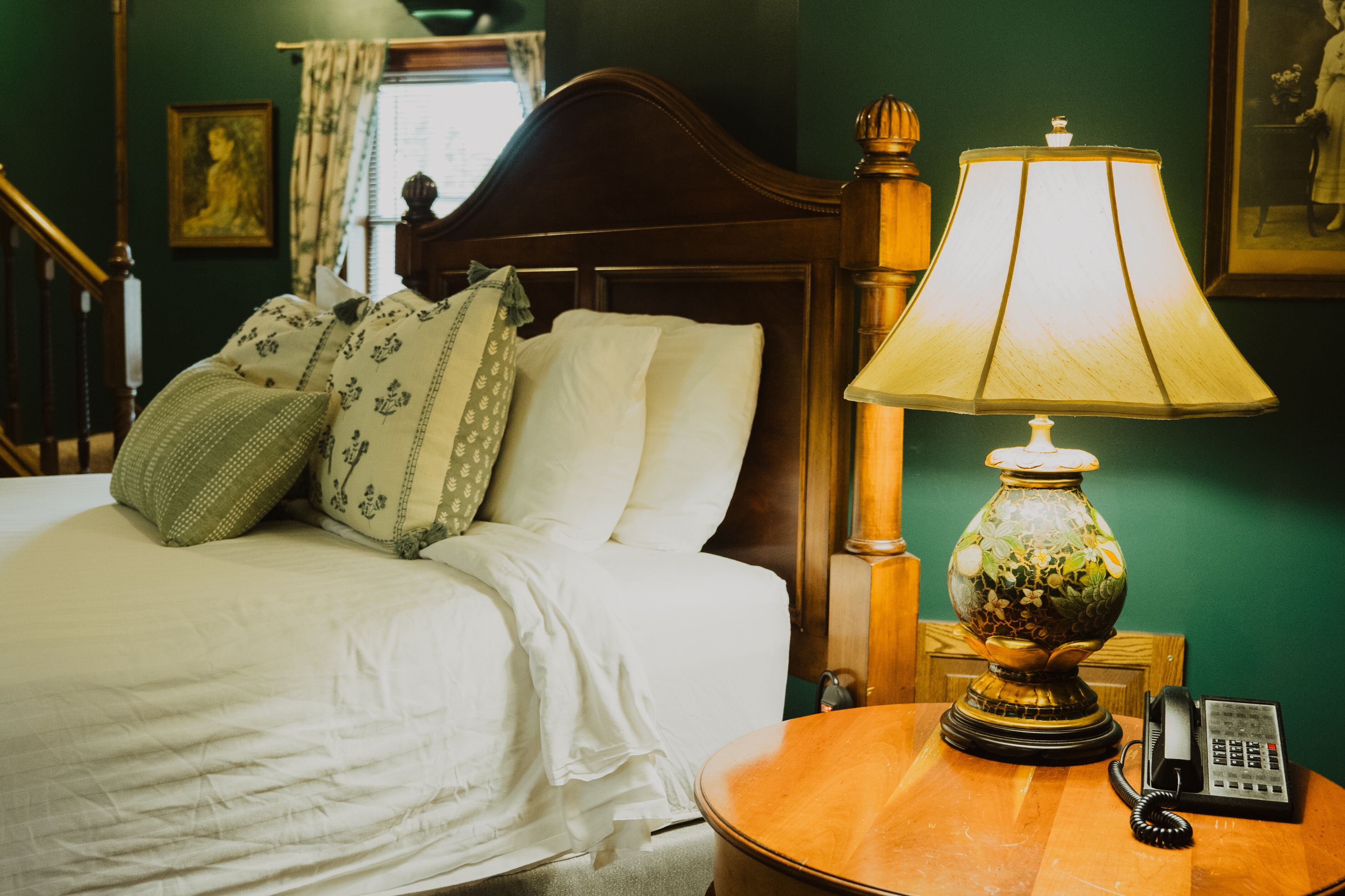 Una cama bien hecha con ropa blanca y almohadones decorativos, una mesita de noche de madera con una lámpara y un teléfono, y una pared verde con un cuadro enmarcado.