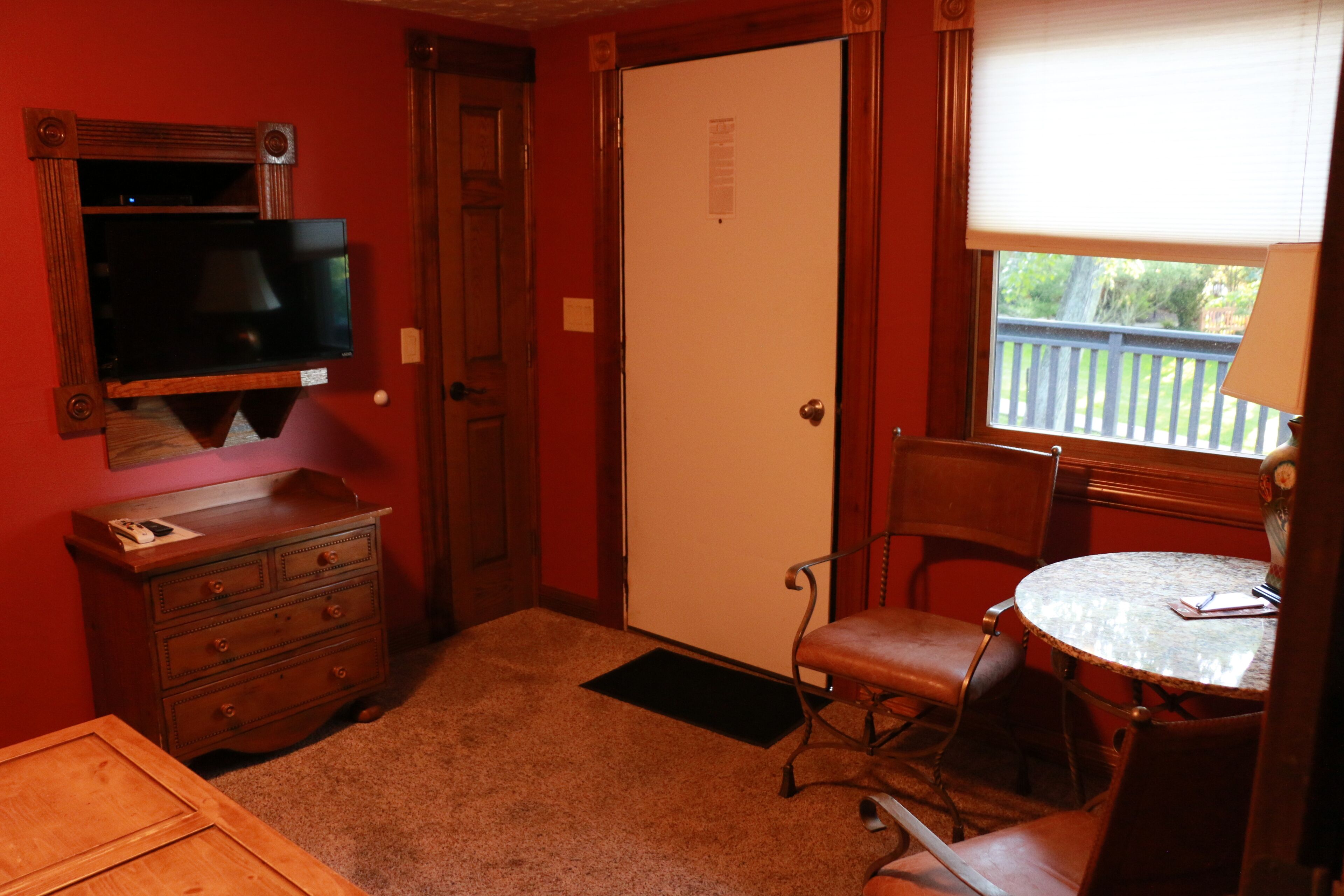 A room with a wooden dresser, a television mounted on the wall, a door, a window with a view of a balcony, and a round table with chairs.