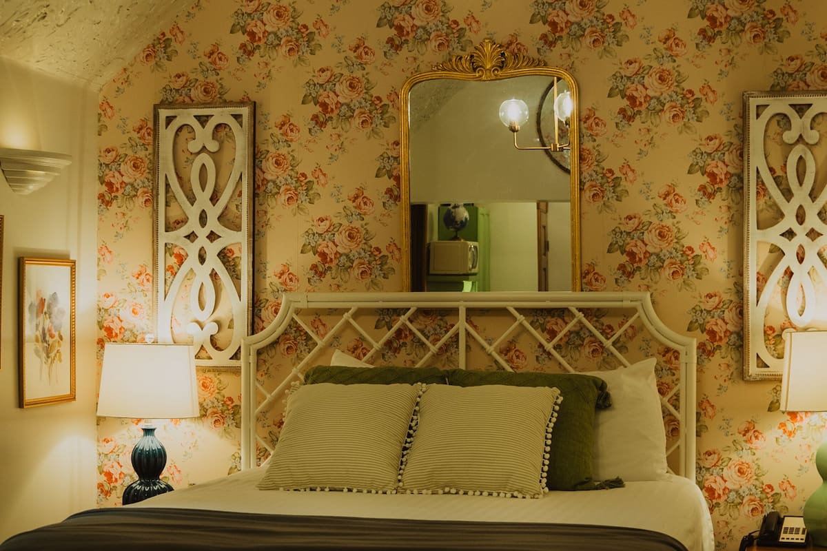 A bedroom with a floral wallpaper, a white bed with a diamond-patterned headboard, a chandelier, and a mirror above the bed.
