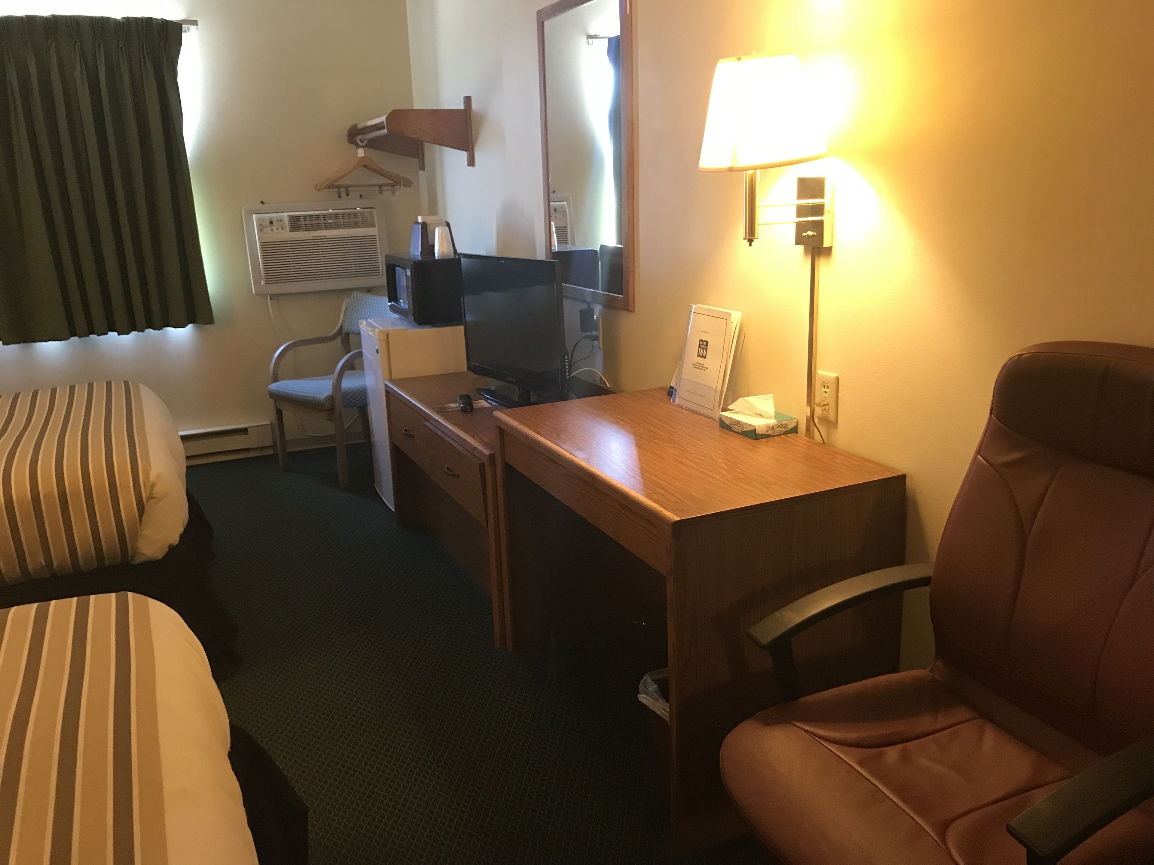 Ein Hotelzimmer mit einem Bett, einem Schreibtisch mit Computer, einem Stuhl, einer Lampe und einem fenster mit Vorhängen.