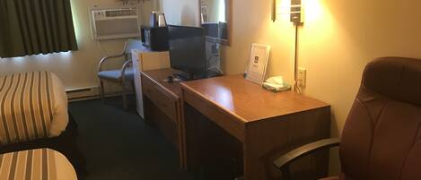 Ein Hotelzimmer mit einem Bett, einem Schreibtisch mit Computer, einem Stuhl, einer Lampe und einem fenster mit Vorhängen.